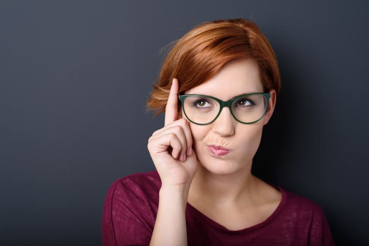 Nerdy scholastic young woman wearing geeky glasses standing thinking with her finger raised and a grimace of concentration in a humorous stereotypical depiction, over a dark background